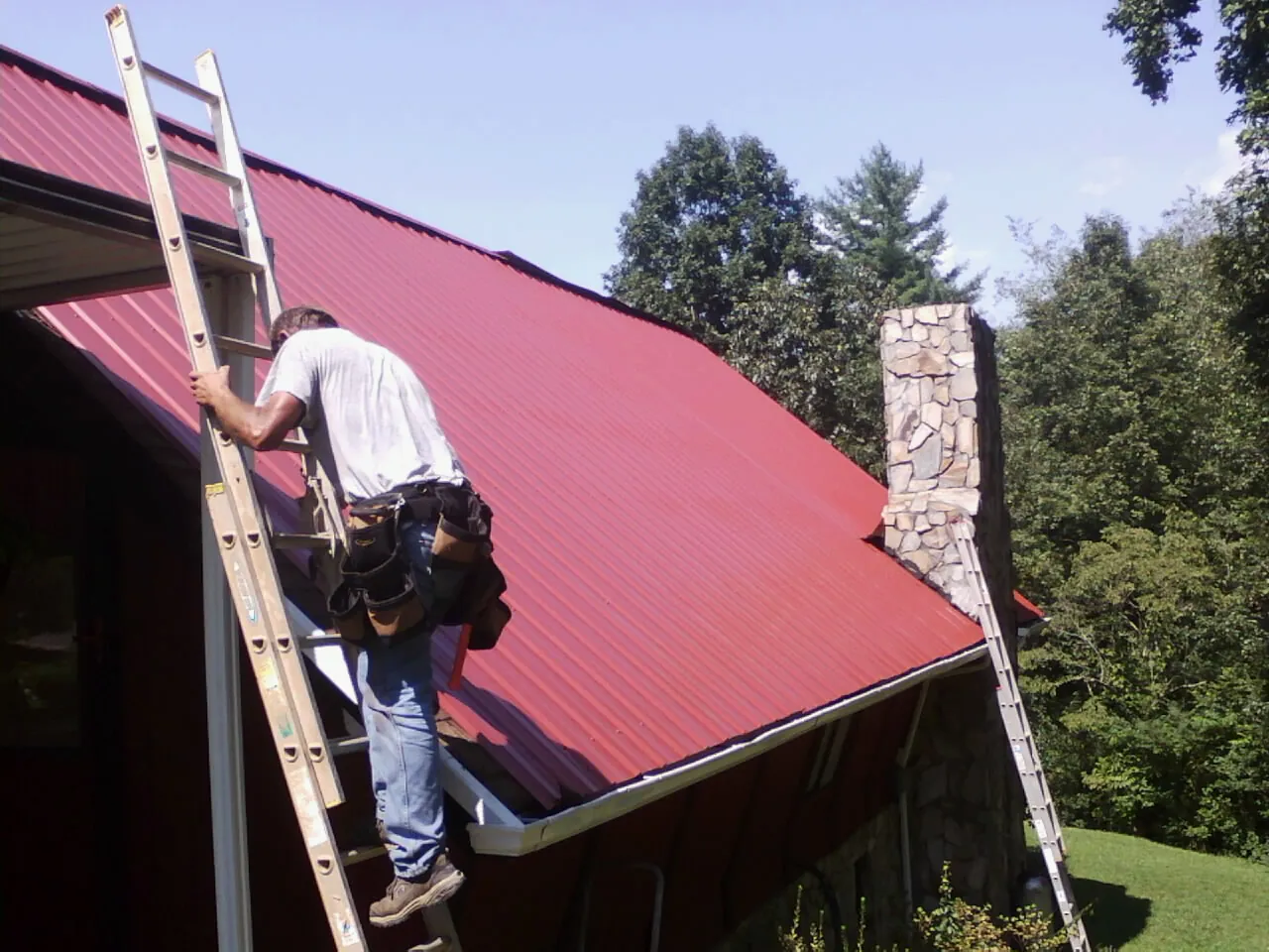 Roofer on a ladder beside a red metal roof and stone chimney — Soffit & Fascia Repair by Roofing Brothers in Buffalo Creek